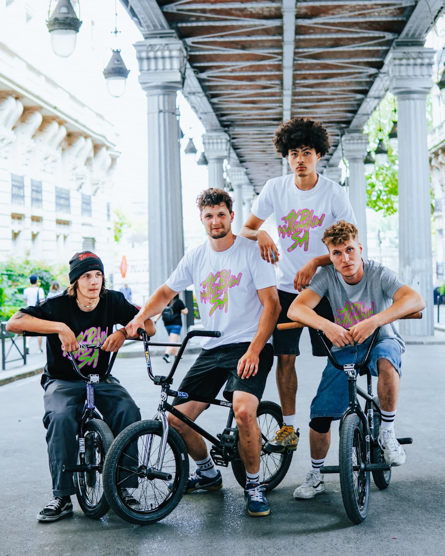 Photo de quatre hommes posant sous le métro aérien de Paris portant des t-shirts ADMS SHOP avec la phrase Ayd Manmanw Sa en rose et vert
