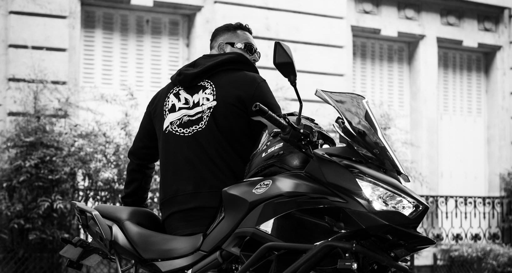 Photo en noir et blanc d'un homme de dos portant un hoodie ADMS SHOP floqué assis sur une moto
