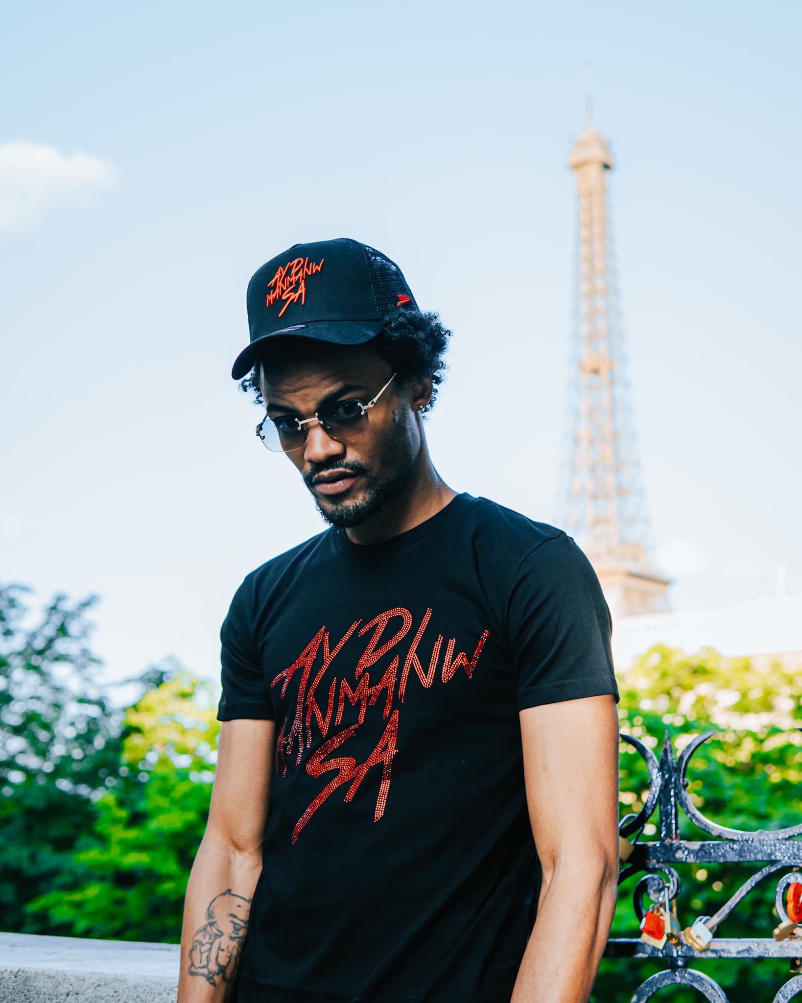 Photo d'un homme posant devant la Tour Eiffel portant un t-shirt et une casquette noir pailleté rouge ADMS SHOP avec le texte Ayd Manmanw Sa